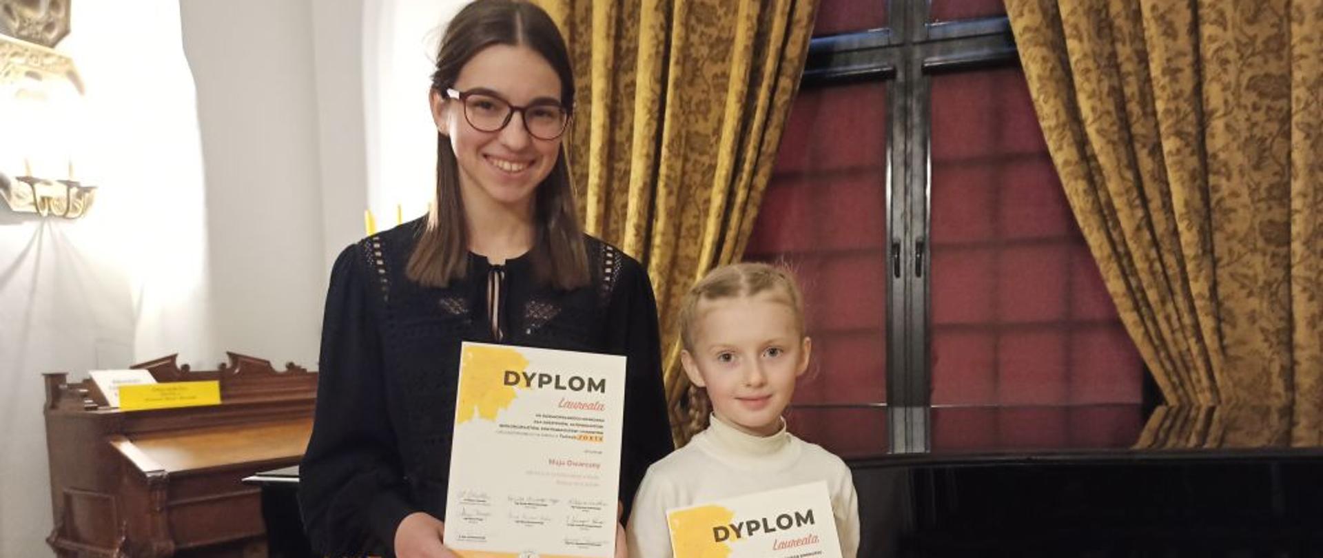 Dwie stojące obok siebie dziewczęta trzymają w rękach Dyplom Laureata VII Ogólnopolskiego Konkursu Forte 2022. Za nimi znajduje się fortepian, a z lewej strony zabytkowa fisharmonia. 