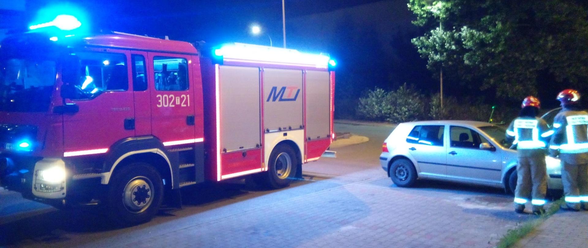 Zdjęcie przedstawia parking gdzie doszło do pożaru samochodu osobowego. Po prawej stronie stoi auto, w którym zapaliła się komora silnika a przy nim dwóch ratowników, po lewej zaś zaparkował wóz strażacki.