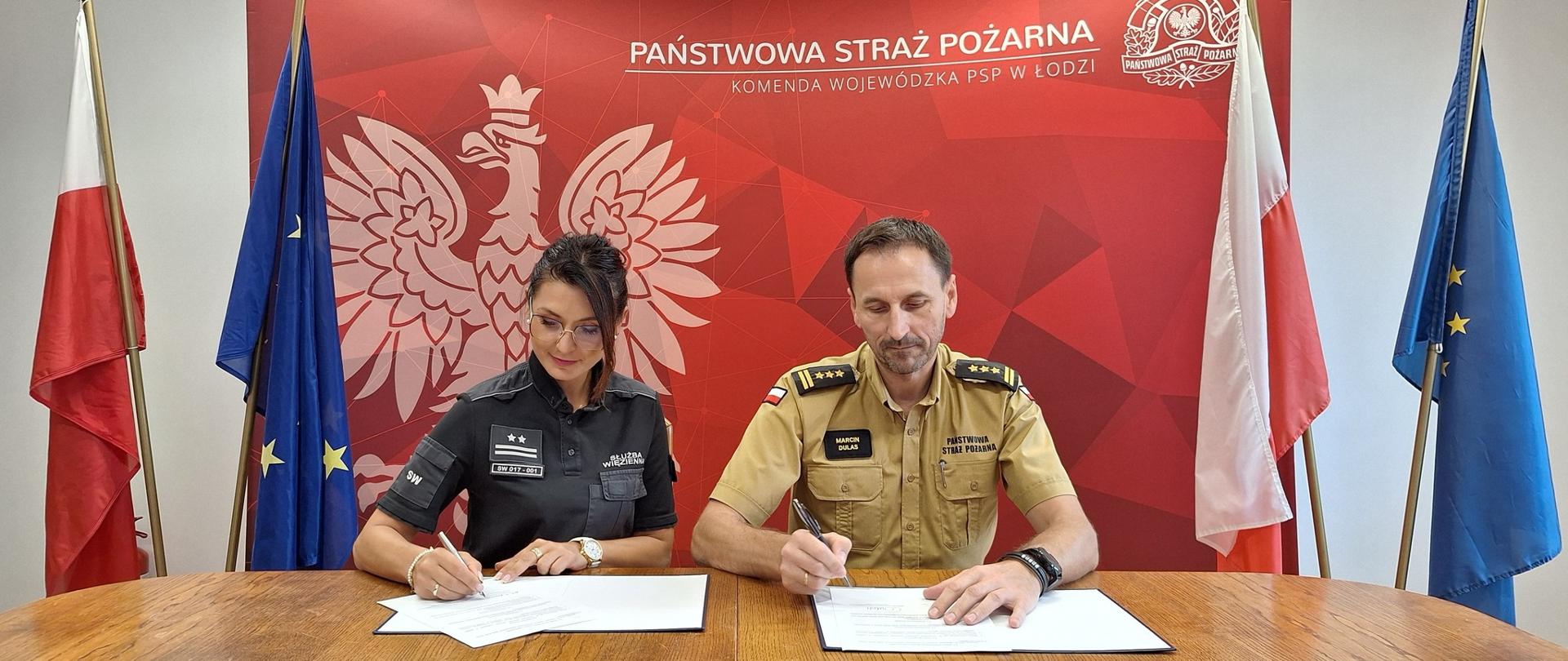 Kobieta w mundurze Służby Więziennej i mężczyzna w mundurze Państwowej Straży Pożarnej siedzą przy stole i podpisują dokumenty. Za nimi widoczne jest czerwone tło z orłem w koronie i napisem „Państwowa Straż Pożarna – Komenda Wojewódzka PSP w Łodzi”. Po bokach ustawione są flagi Polski i Unii Europejskiej, a nad nimi zawieszony jest herb Polski.