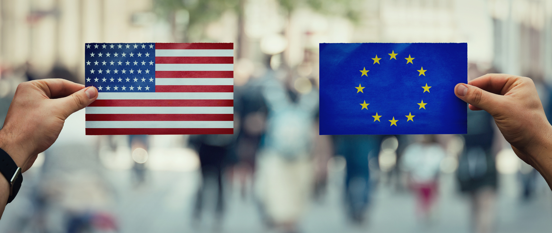 Flagi USA i Unii Europejskiej trzymane w dłoniach. W tle widok ulicy (zamazany).