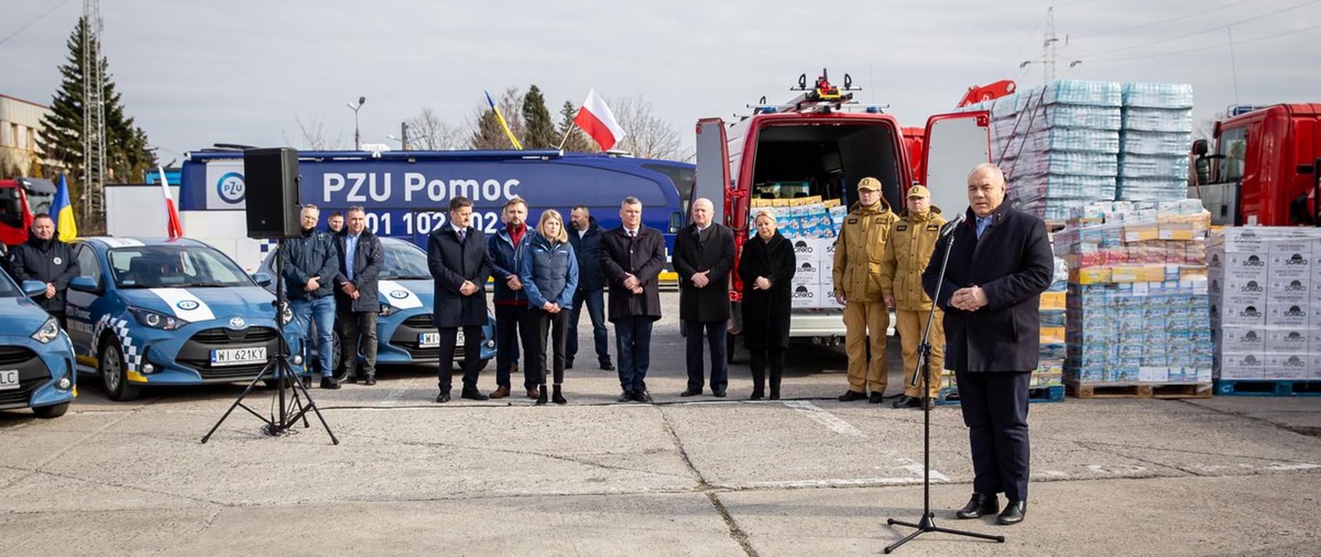 Wicepremier Jacek Sasin stoi przy mikrofonie. W tle samochody firmowe PZU oraz dostawczaki z suchym prowiantem i wodą, produkty również na paletach na placu. Za premierem stoją przedstawiciele spółek i samorządu. 