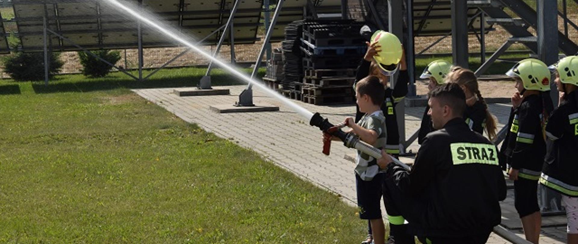 Grupa przedszkolaków podczas próby gaszenia symulowanego pożaru. Zajęcia prowadzi asp. Wojciech Dymowski.