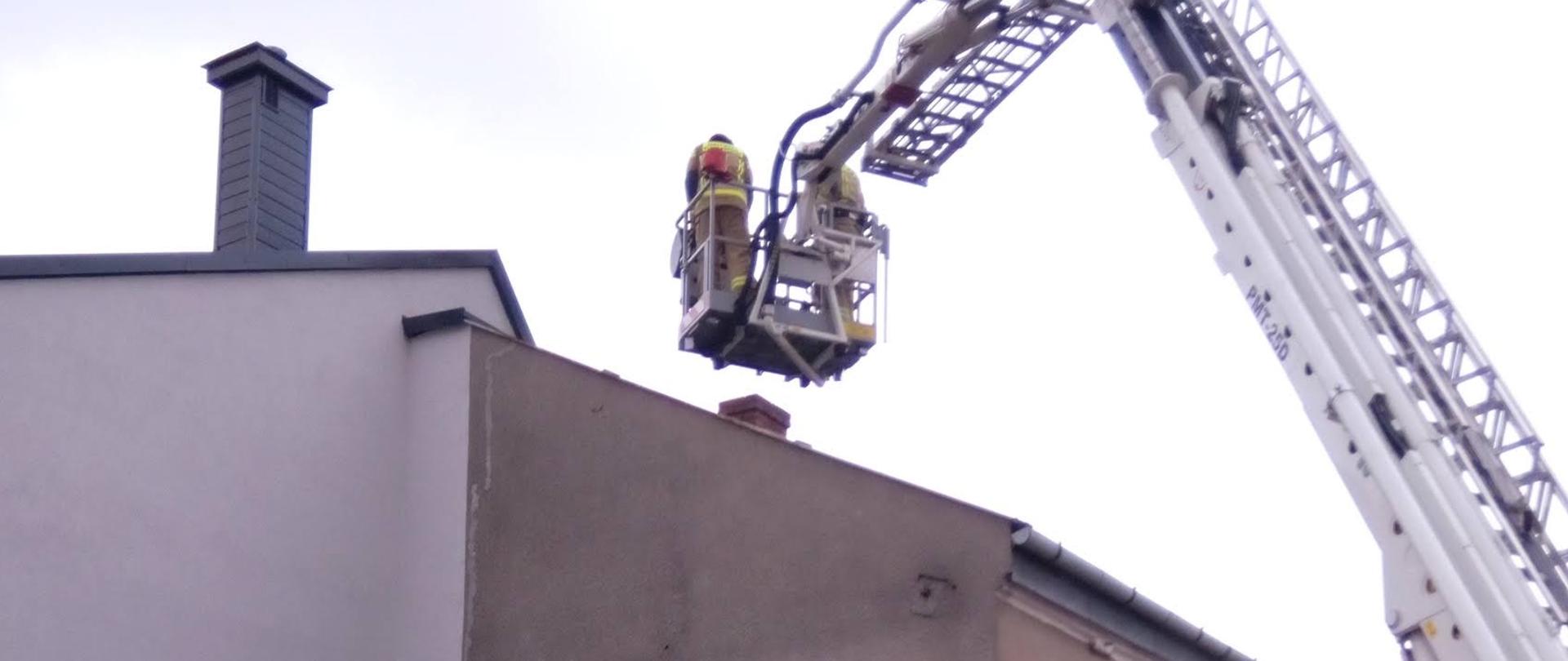 Zdjęcie przedstawia podnośnik hydrauliczy wraz z trzema strażakami, dwóch znajdujących się w koszu oraz jednego operatora podnośnika. Podnośnik hydrauliczny stoi przed budynkiem mieszkalnym 