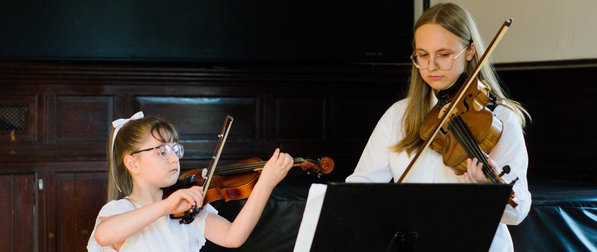 Dwie skrzypaczki podczas koncertu. Jest to duet złożony z dziewczynki oraz nastolatki. Obie ubrane są w białe stroje. Grają na skrzypcach z uwagą patrząc na nuty ustawione na pulpicie. W tle widać fragment fortepianu.