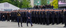 Prezydent RP przechodzi przed zwartym szykiem funkcjonariuszy PSP w galowych mundurach; oficer PSP salutuje, a w tle widać licznych fotografów i operatorów kamer dokumentujących wydarzenie.