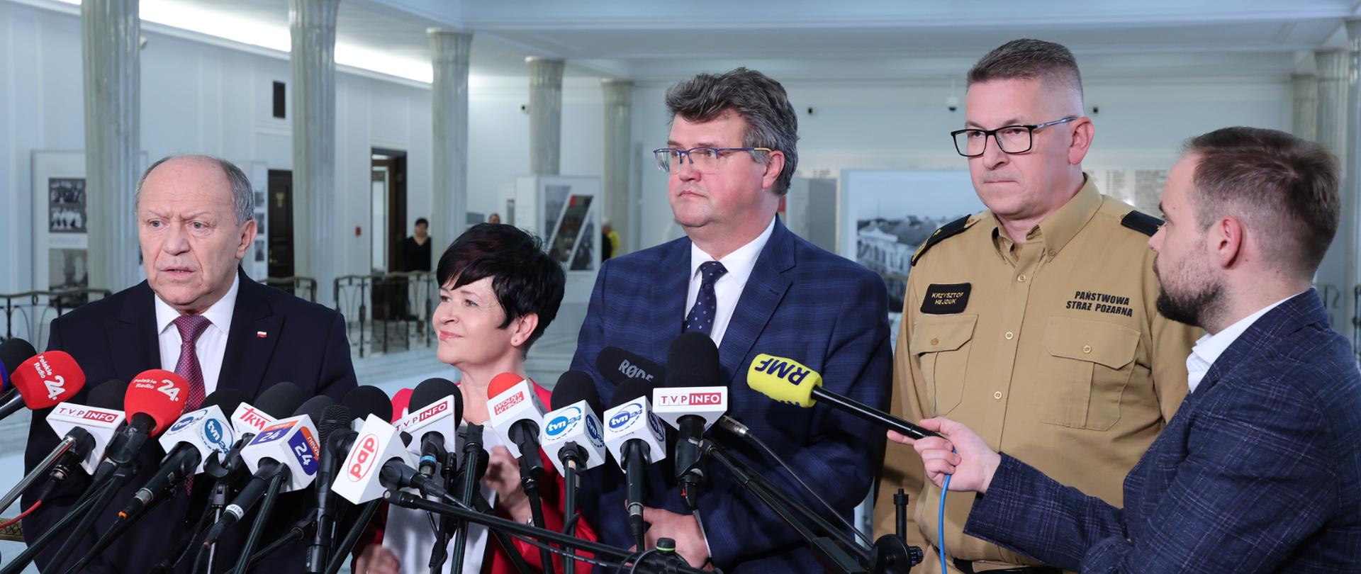 Na zdjęciu przedstawiciele MSWiA oraz Zastępca Komendanta Głównego PSP podczas konferencji dot. dotacji dla MDP. Po prawej stronie kadru dziennikarz radiowy trzymający mikrofon.