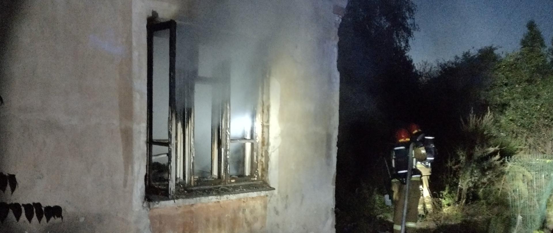 Przed budynkiem z którego wydobywa się dym z otwartego okna stoi dwóch strażaków, którzy dogaszają pożar jaki wybuchł w kuchni.