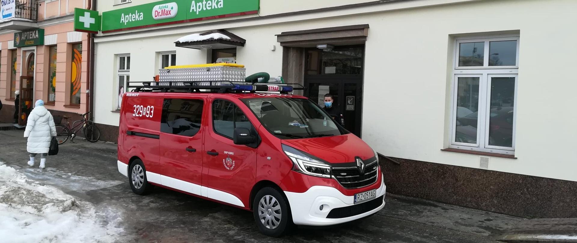 Na zdjęciu w centralnej części przed budynkiem wielorodzinnym stoi samochód typu bus w kolorze czerwonym. Budynek dwukondygnacyjny z poddaszem, na zdjęciu widoczne dwie kondygnacje. Tuż za budynkiem wejście do obiektu. Obok samochodu stoi strażak OSP, w lewej części zdjęcia widoczny przechodzień odwrócony tyłem. Po lewej stronie budynku na zdjęciu widoczne dwie apteki. 