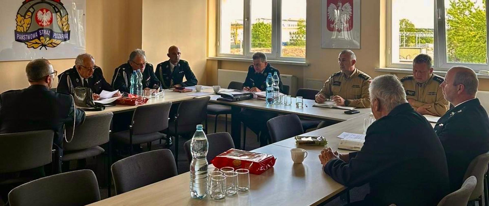 Sala konferencyjna podczas spotkania służbowego strażaków. Kilku mężczyzn w mundurach siedzi przy ustawionych w podkowę stołach, prowadząc naradę. Na ścianie widoczne jest godło Państwowej Straży Pożarnej, a w pomieszczeniu znajdują się okna, projektor sufitowy oraz butelki z wodą i dokumenty na stołach.