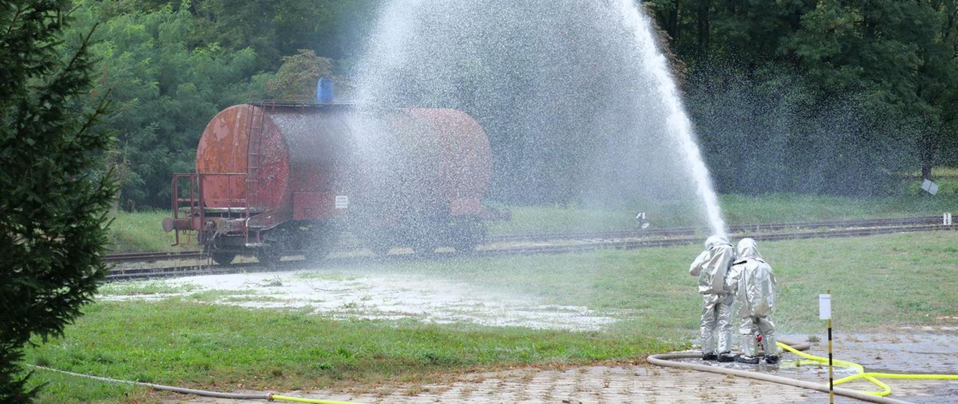 W trakcie ćwiczeń pod kryptonimem "BAZA 22" dwóch strażaków w ubraniach żaroodpornych podaje pianę ciężką na cysternę kolejową.