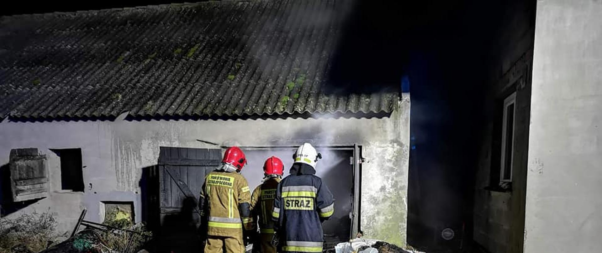 Pora nocna, dwóch strażaków PSP w piaskowych ubraniach i czerwonym hełmie oraz strażak OSP w granatowym ubraniau i białym hełmie stoją przed budynkiem z którego wydobywa się szary dym i para wodna. Na ziemi leży żółta porozrzucana słoma Miejsce zdarzenie oświetlone z reflektora.