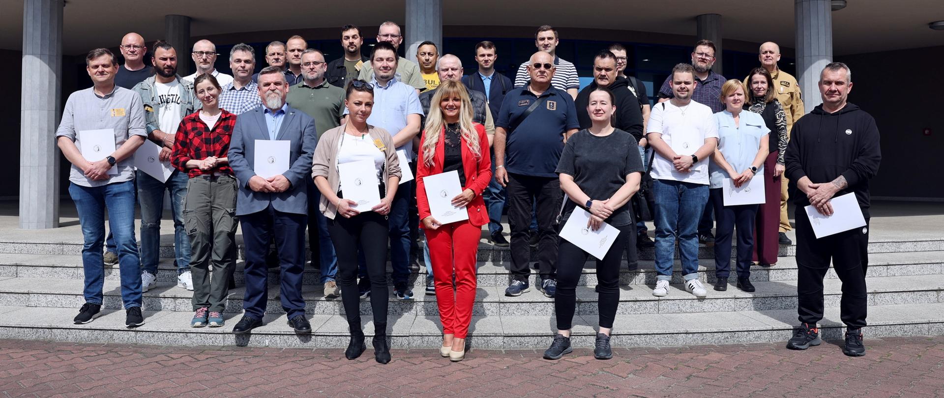 Pamiątkowe zdjęcie grupowe uczestników szkolenia inspektorów ochrony przeciwpożarowej (27 osób) w towarzystwie oficera CS PSP oraz kobiety