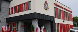 Widok z zewnątrz na nowoczesny budynek Państwowej Straży Pożarnej z czerwonymi elementami elewacji. Przy wejściu ustawiono biało-czerwone flagi, pamiątkowy dzwon.