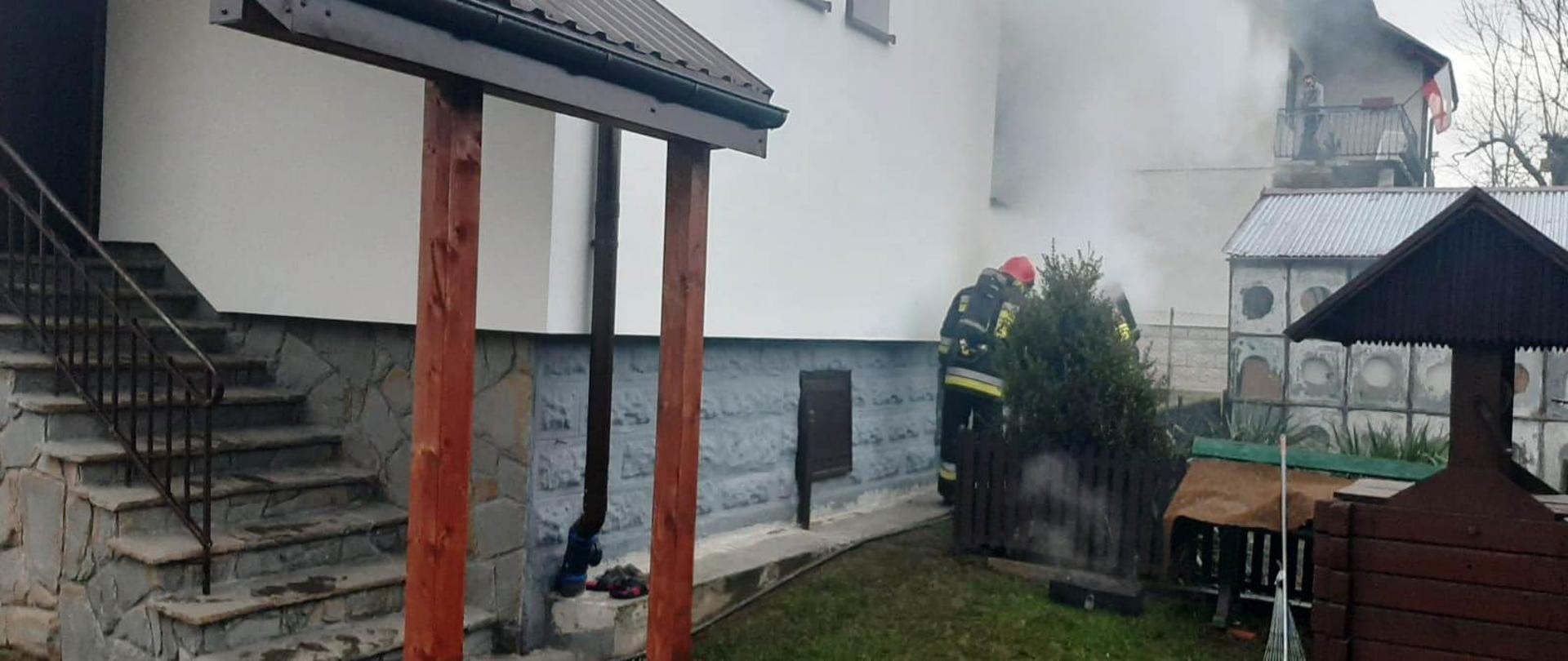 Dym wydobywający się z kotłowni budynku mieszkalnego oraz strażacy prowadzący działania.