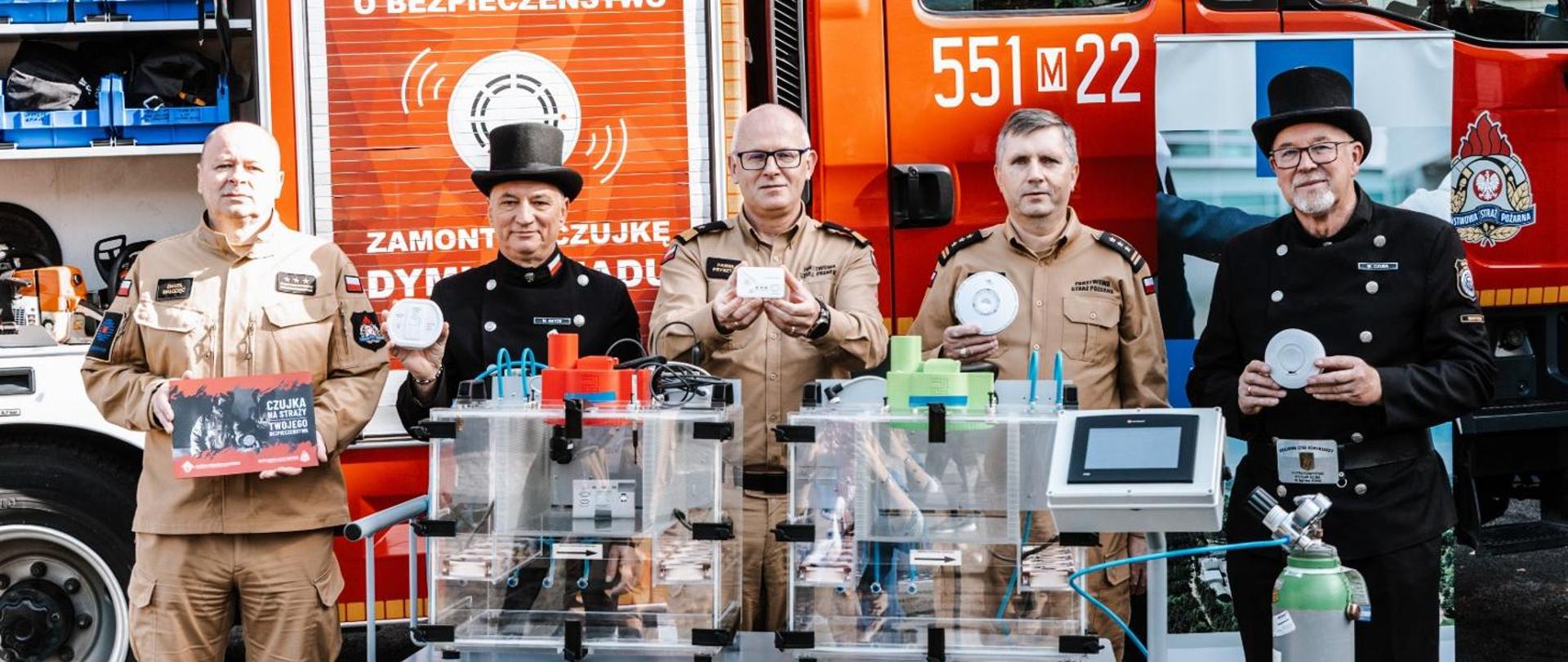 Na zdjęciu widać trzech oficerów Państwowej Straży Pożarnej w mundurach służbowych oraz dwóch kominiarzy propagujących akcję czujka na straży twojego bezpieczeństwa. Osoby te stoją na tle samochodu pożarniczego 