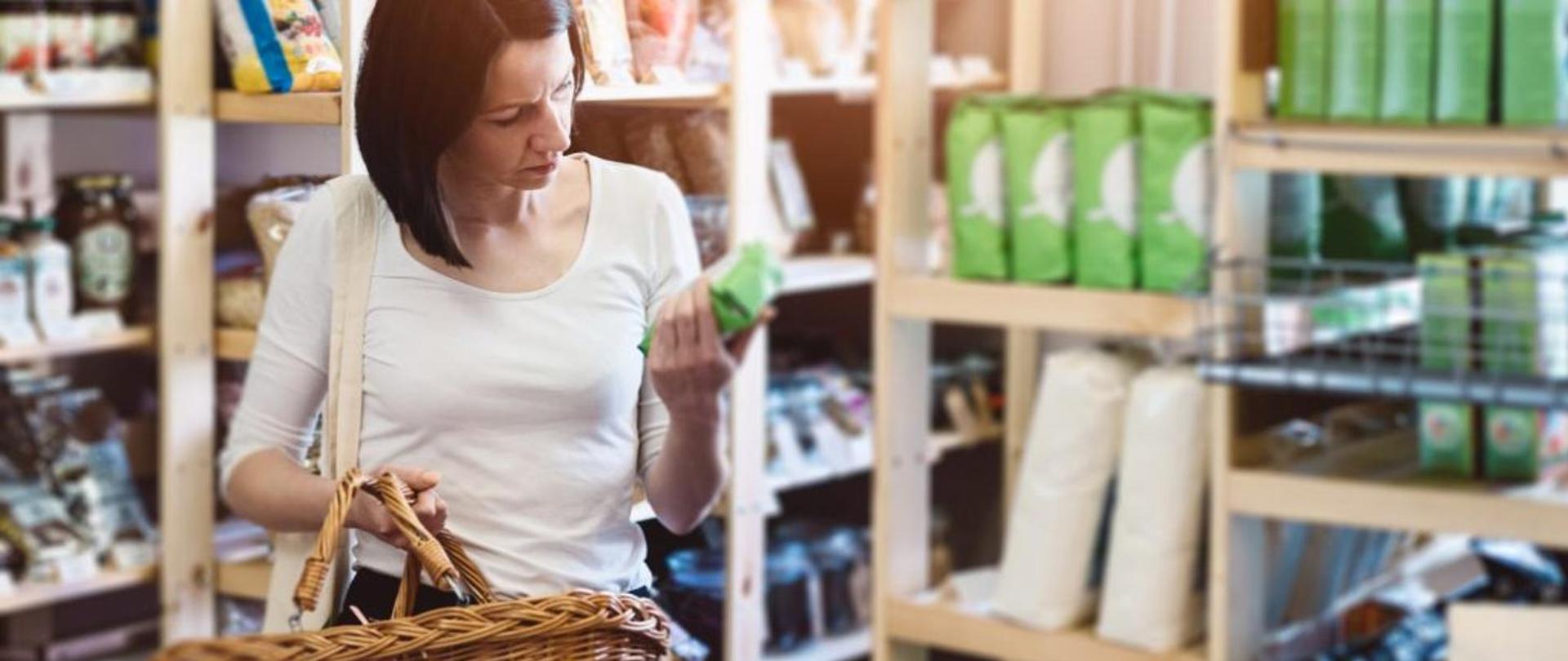 Kobieta robiąca zakupy w sklepie samoobsługowym. W prawej ręce trzyma wiklinowy kosz. W tle rozmyte drewniane półki sklepowe.