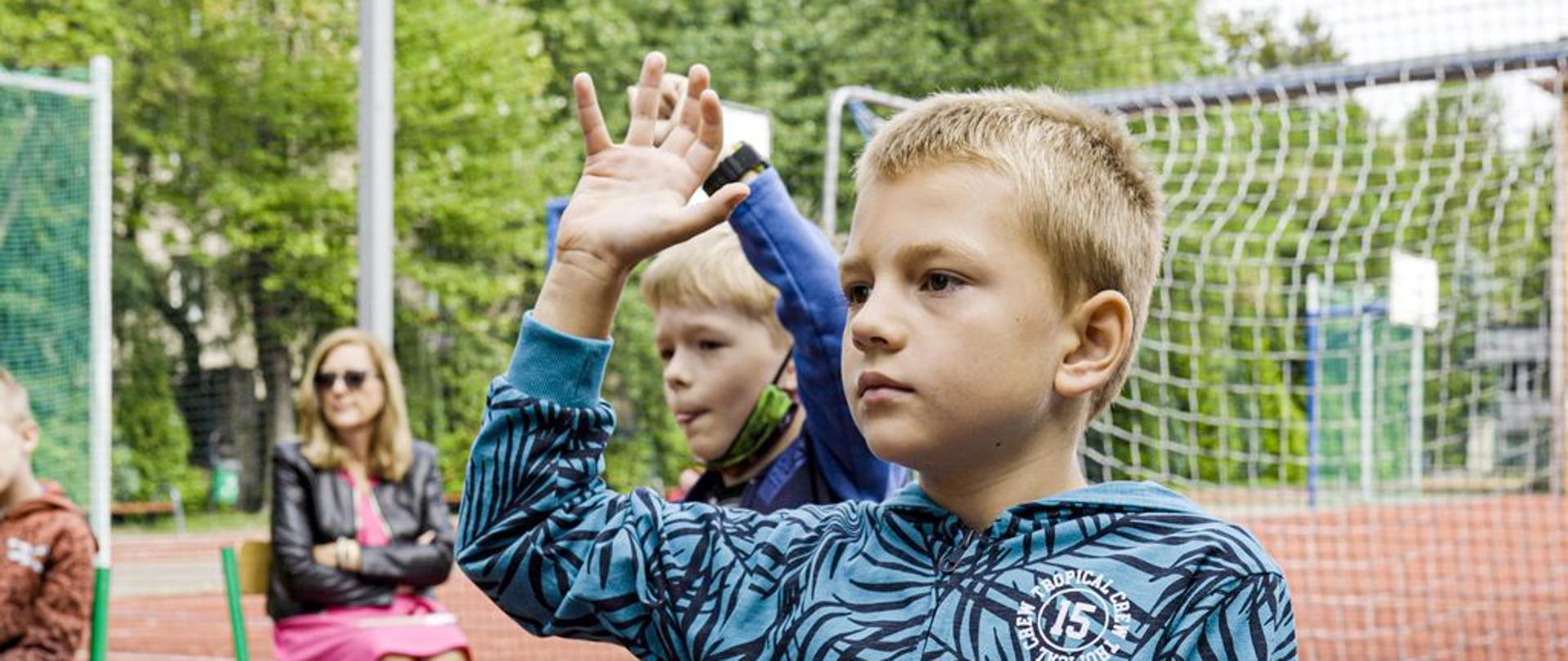 Na pierwszym planie siedzi chłopiec - uczestnik warsztatów, z uniesioną do góry ręką, zgłaszający się do odpowiedzi, za nim w tle kolejny uczestnik, osoba dorosła oraz boisko szkolne, na którym odbywały się warsztaty. 