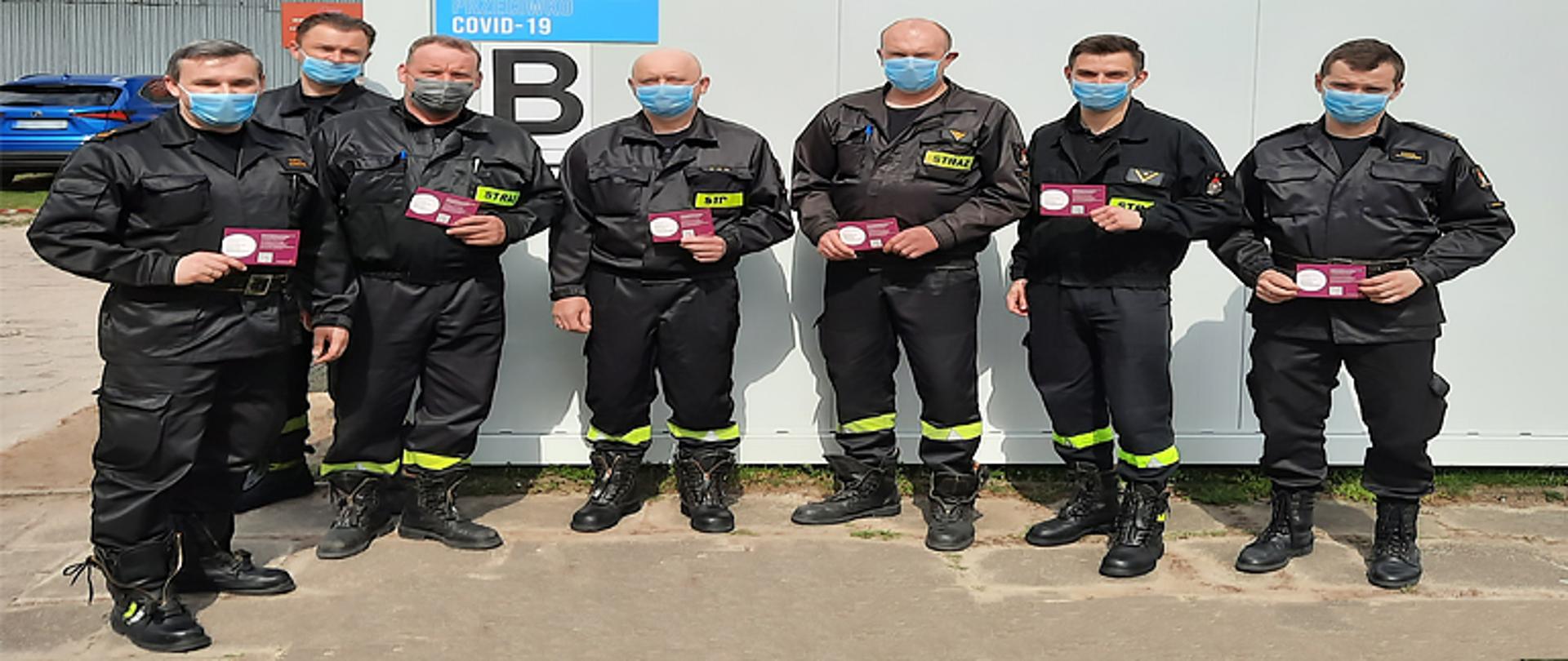 Zdjęcie przedstawia sześciu strażaków z Jednostki Ratowniczo-Gaśniczej w Łowiczu oraz zastępcę komendanta powiatowego tuż po zaszczepieniu przeciw COVID-19 trzymających w ręku zaświadczenie potwierdzające zaszczepienie przeciwko wirusowi Sars-Cov-2. Ubrani są w ubrania koszarowe koloru czarnego, na twarzy mają maseczkę, za nimi budynek koloru szarego z napisem Punkt szczepień przeciwko Covid-19 z dużą widoczną literą B, w tle budynek administracyjny.