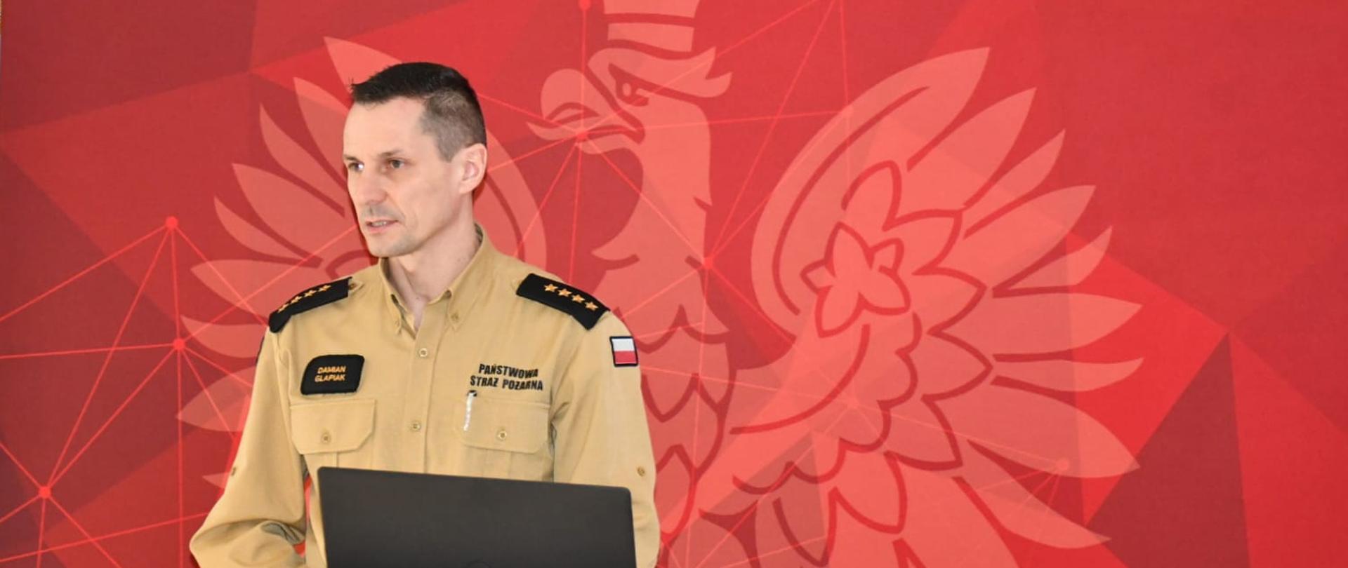 Zdjęcie przedstawia oficera Państwowej Straży Pożarnej w Polsce, który stoi przy podium i prowadzi prezentację lub przemówienie. Ma na sobie mundur wyjściowy w kolorze khaki z naszywką polskiej flagi na ramieniu oraz nazwiskiem na piersi. Na jego pagonach widnieją cztery gwiazdki, co sugeruje stopień oficerski.
Za nim znajduje się czerwone tło z dużym, stylizowanym wizerunkiem orła oraz napisem "PAŃSTWOWA STRAŻ POŻARNA" i "KOMENDA POWIATOWA PAŃSTWOWEJ STRAŻY POŻARNEJ W WOLSZTYNIE", co wskazuje, że zdjęcie zostało wykonane w siedzibie jednostki PSP w Wolsztynie. W prawym górnym rogu widoczne jest również logo PSP.
Przed strażakiem znajduje się laptop podłączony kablem, co sugeruje, że prowadzi prezentację multimedialną lub odprawę dla innych funkcjonariuszy. Jego postawa oraz mimika wskazują na skupienie i zaangażowanie w wystąpienie.
