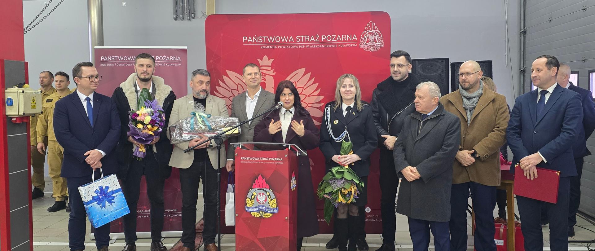 Wokół mównicy zgromadzeni ludzie, kilkanaście osób. Jedna przedstawicielka grupy przemawia. Wielu z nich trzyma kwiaty