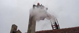 Na pierwszym planie zdjęcia widać budynek z wysokim kominem, w którym doszło do zapalanie sadzy. Z komina wydobywają się kłęby jasnego dymu, a tuż za nimi można dostrzec strażaków, który pracują w koszu podnośnika hydraulicznego usuwając zagrożenie. 