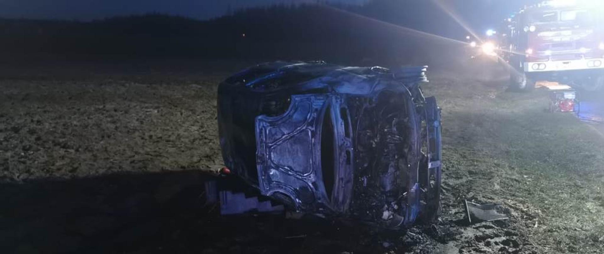 Na pierwszym planie zdjęcia poza drogą leży na boku wrak spalonego pojazdu, na drugim planie samochód ratowniczo-gaśniczy oraz strażacy w ubraniach specjalnych.