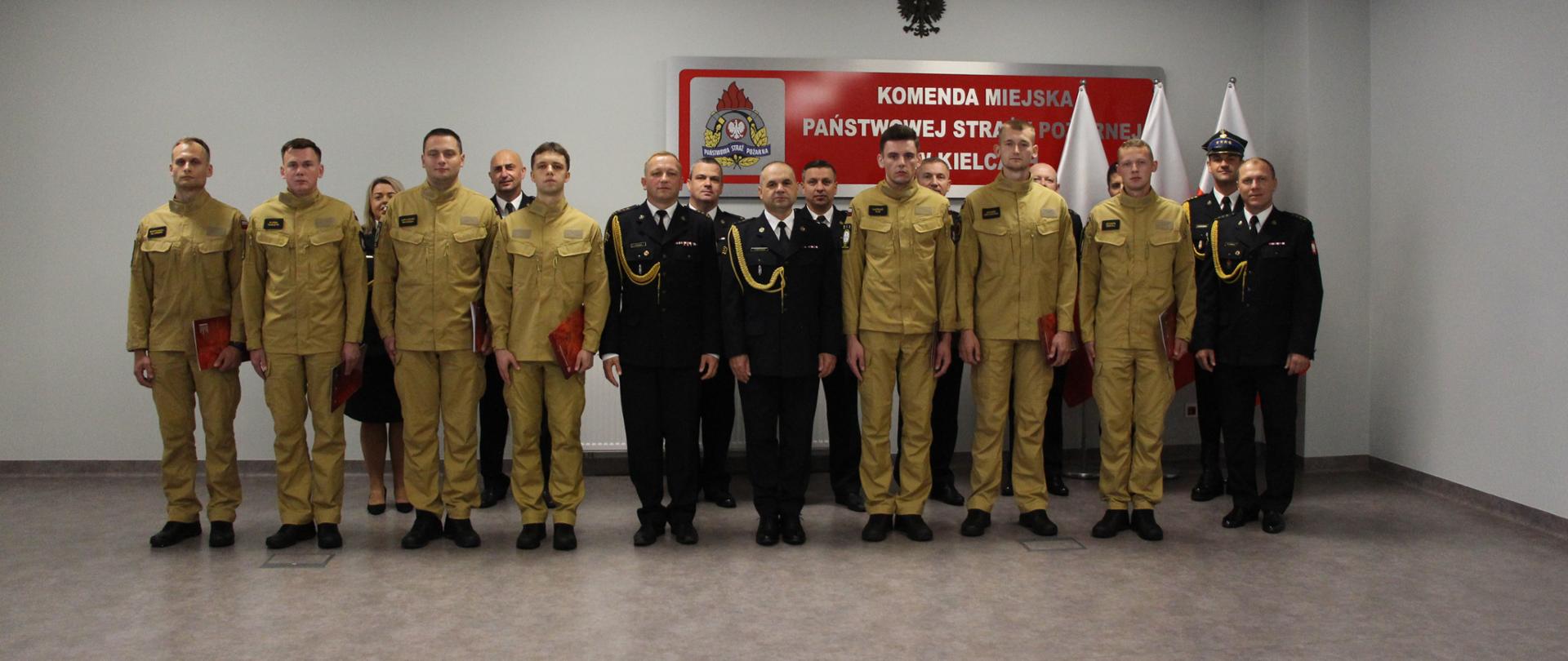 Na zdjęciu stoją ustawieni w pierwszym szeregu nowo przyjęci strażacy wraz zastępcami komendanta miejskiego Państwowej Straży Pożarnej w Kielcach i naczelnikiem wydziału organizacyjno kadrowego. W drugim rzędzie stoją dowódcy jednostek oraz naczelnicy wydziałów tutejszej komendy.