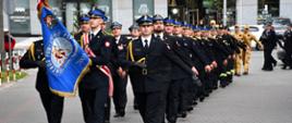 Pododdział strażaków mundurach galowych podczas defilady maszeruje równym krokiem po placu miejskim. Na czele, po lewej stronie, idzie poczet sztandarowy – strażak w środku niesie duży, niebieski sztandar z napisem „KOMENDA POWIATOWA PSP W MIŃSKU MAZOWIECKIM 2003” i wizerunkiem świętego Floriana. Dłonie maszerujących strażaków w czarnych rękawiczkach są wyciągnięte do boku w marszowym geście.