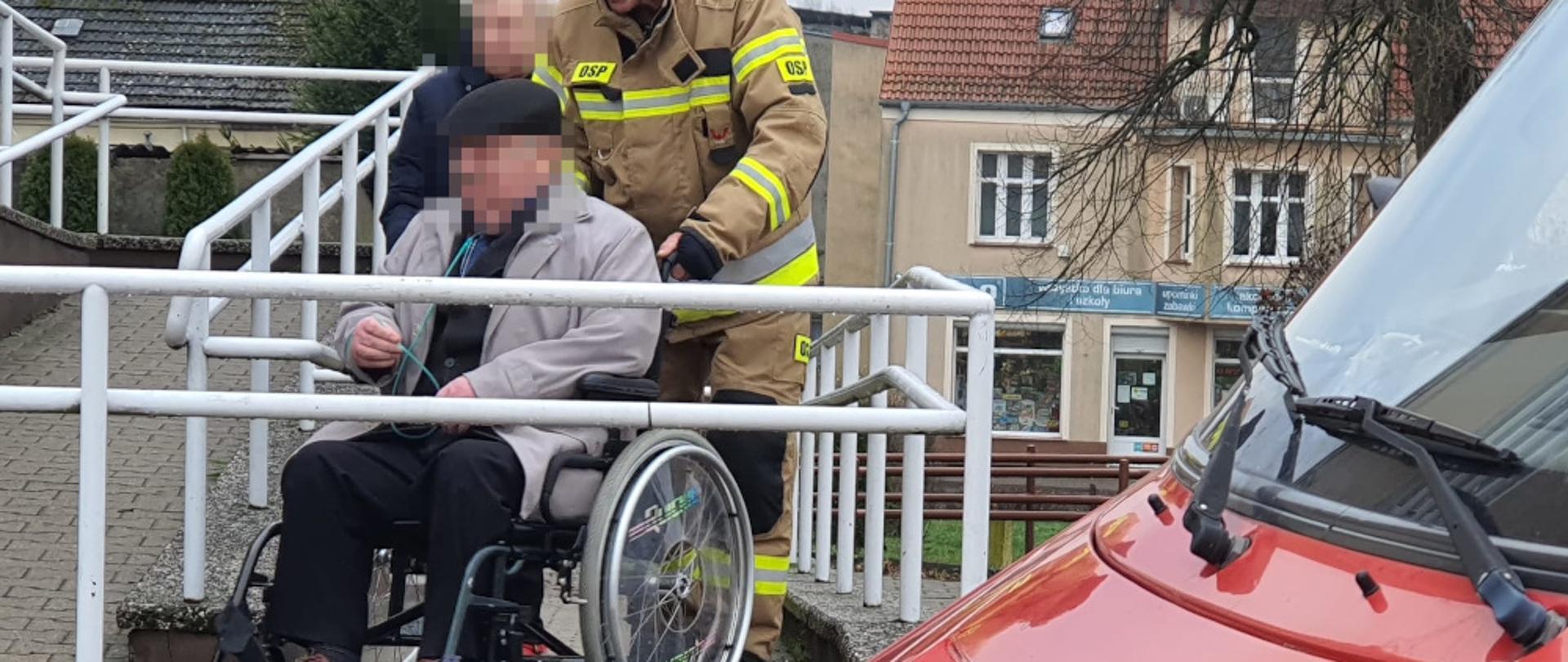 Strażak z OSP Barwice dh Mirosław Tojnacki ubrany w ubranie specjalne, koloru pisakowego z napisami OSP na podjeździe dla niepełnosprawnych, transportuje osobę niepełnosprawną (mężczyznę w starszym wieku) na wózku inwalidzkim. Obok idzie kobieta ubrana w kurtkę granatową i niesie butlę z tlenem niezbędną do oddychania osobie na wózku.