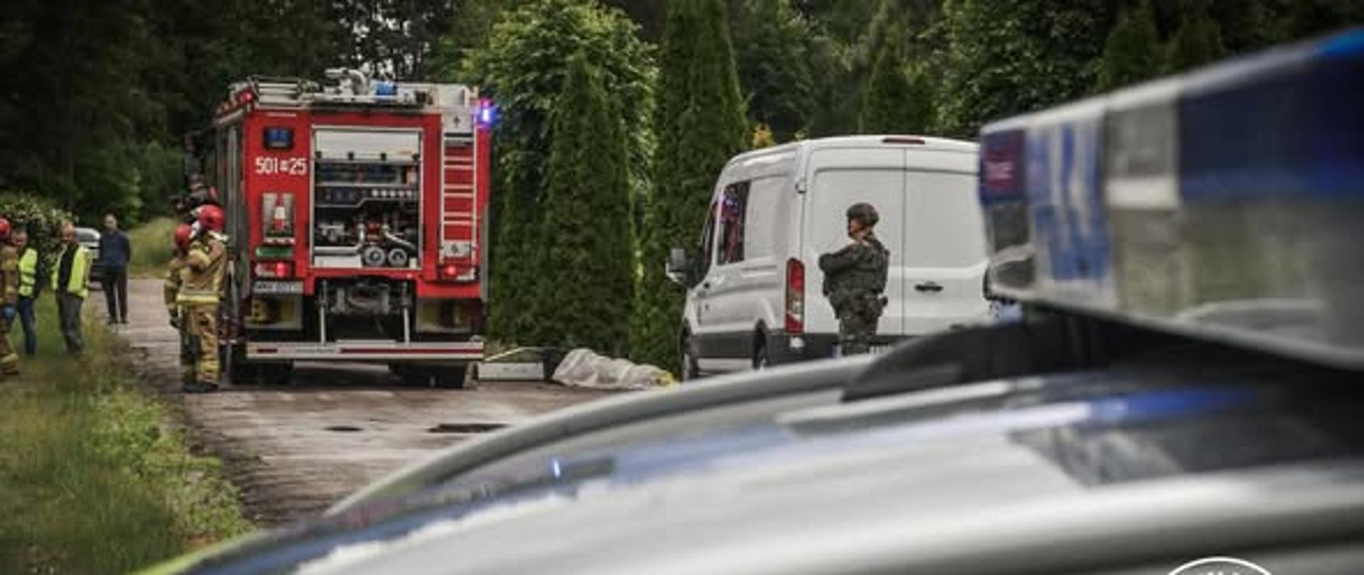 Na zdjęciu widać wóz strażacki, przy którym stoją funkcjonariusze PSP, z prawej strony stoi biały bus a przy nim stoi żołnierz. Przez środek biegnie droga a dookoła są drzewa