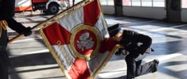 Oficer Państwowej Straży Pożarnej przyklęka i całuje płat sztandaru.