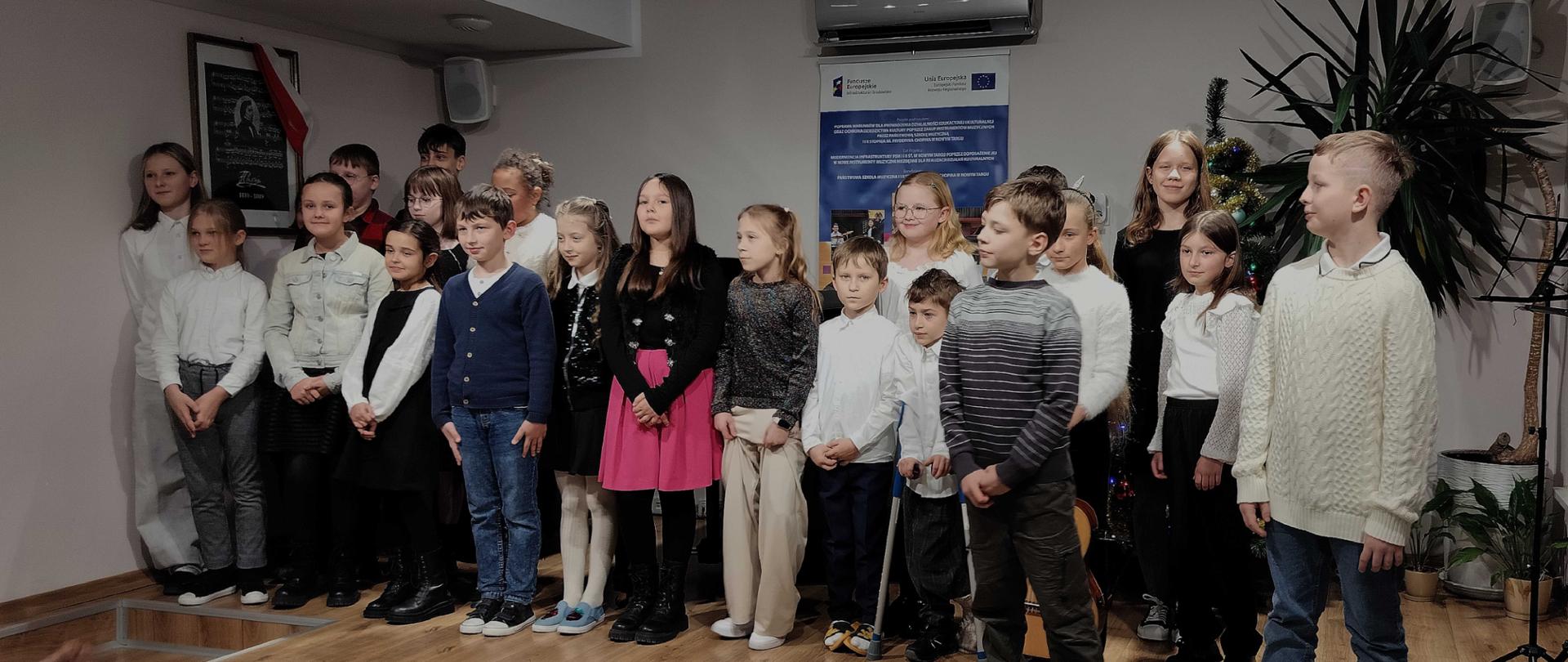 Zdjęcie przedstawia grupę dzieci ustawionych w kilku rzędach na niewielkiej, drewnianej scenie w pomieszczeniu wewnętrznym. Widać ich około 20, w wieku szkolnym, zarówno dziewczęta, jak i chłopców. Stoją blisko siebie, niemal ramię w ramię, patrząc w stronę osoby, która robi zdjęcie lub ewentualnie w stronę widowni. Wielu ma uśmiechy lub neutralne wyrazy twarzy; wyglądają na gotowych do występu lub właśnie po występie. Ubiór jest raczej oficjalny lub półformalny: kilka dziewcząt ma sukienki albo spódnice, jedna nosi wyraźnie różową sukienkę; inne dziewczęta i chłopcy mają białe koszule, swetry, marynarki, ciemne spodnie, czasem jasne lub kremowe elementy; obuwie różne: od czarnych butów, przez botki, po sportowe buty; niektórzy noszą ciemne skarpetki lub rajstopy. Scena ma drewnianą podłogę i stopnie po lewej stronie, którymi można zejść na poziom widowni. W tle, za dziećmi, wisi na ścianie niewielka, prostokątna tablica lub plakat z tekstem i elementami graficznymi, a wyżej nad nim zamontowana jest jednostka klimatyzacji ściennej. Po prawej stronie widać dużą roślinę doniczkową oraz prawdopodobnie choinkę albo inną dekorację zagożoną świątecznie, co sugeruje sezonowe wydarzenie. Po lewej stronie, na ścianie, znajduje się ramka z obrazem lub fotografią, a pod nią coś w rodzaju głośnika lub innego urządzenia. Oświetlenie: sufit jest niski, z kilkoma okrągłymi, wpuszczonymi lub półwpuszczonymi oprawami świetlnymi, które rzucają równomierne światło na scenę. Całe pomieszczenie ma spokojny, wnętrzarski charakter, jak sala mała lub średniej wielkości, używana do prezentacji, koncertów szkolnych czy spotkań. Ogólny nastrój: grupa wygląda na zorganizowaną i spokojną, jakby trwała chwila formalnego zdjęcia zbiorowego podczas szkolnego lub lokalnego wydarzenia. Zdjęcie ukazuje różnorodność dzieci, ich strojów i niewielką scenę z prostym, ale funkcjonalnym otoczeniem.