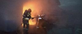 Zdjęcie przedstawia dwóch strażaków prowadzących działania gaśnicze podczas pożaru garażu i samochodu (widoczny ognień wydobywający się z budynku)