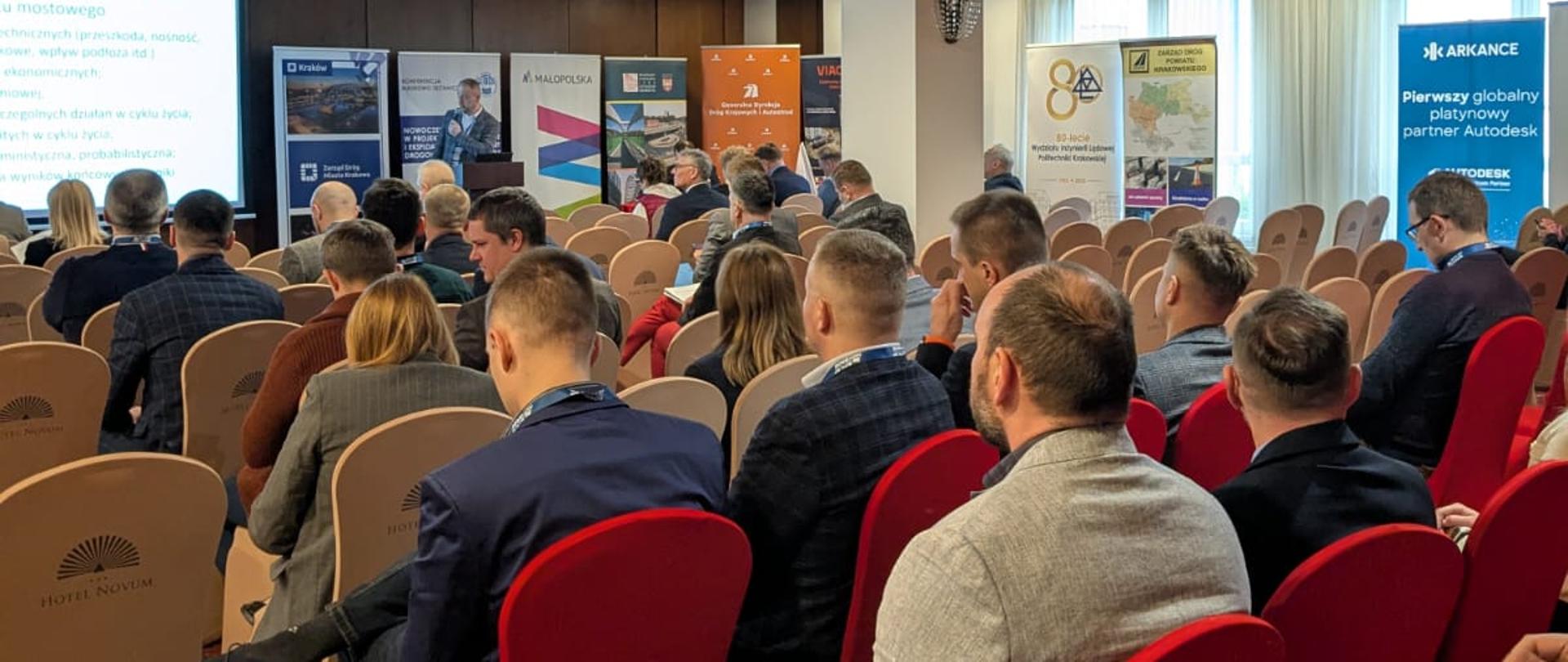 Rozpoczęcie drugiego dnia konferencji Novdrog w Hotelu Novum w Niepołomicach. Na sali uczestnicy siedzący na krzesłach w układzie teatralnym. Widoczne wystąpienie jednego z prelegentów.