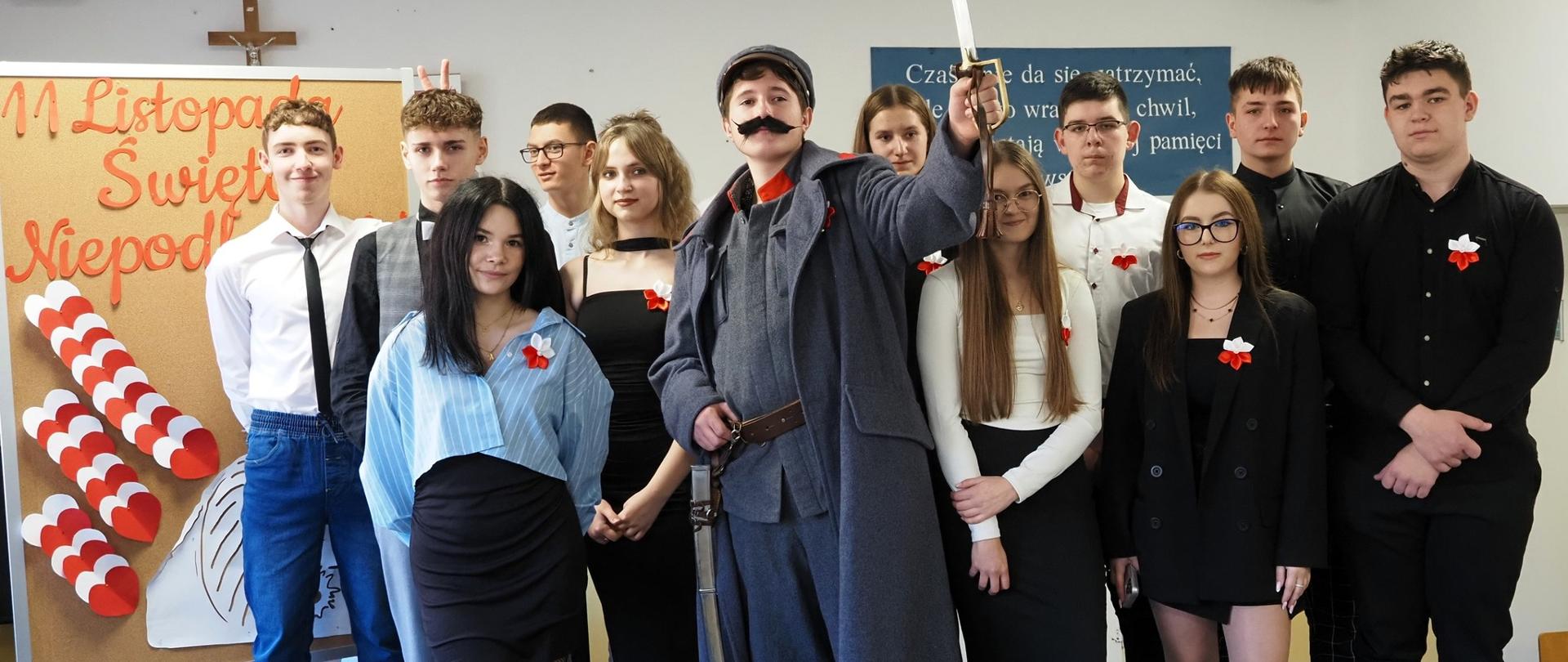 Zdjęcie grupowe wykonawców akademii: pośrodku stoi „Piłsudski” z uniesioną szablą, wokół niego uczniowie w eleganckich strojach z biało-czerwonymi kotylionami. W tle tablica z napisem „11 Listopada Święto Niepodległości” – wyraźne zdjęcie pamiątkowe całej ekipy.