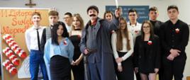 Zdjęcie grupowe wykonawców akademii: pośrodku stoi „Piłsudski” z uniesioną szablą, wokół niego uczniowie w eleganckich strojach z biało-czerwonymi kotylionami. W tle tablica z napisem „11 Listopada Święto Niepodległości” – wyraźne zdjęcie pamiątkowe całej ekipy.