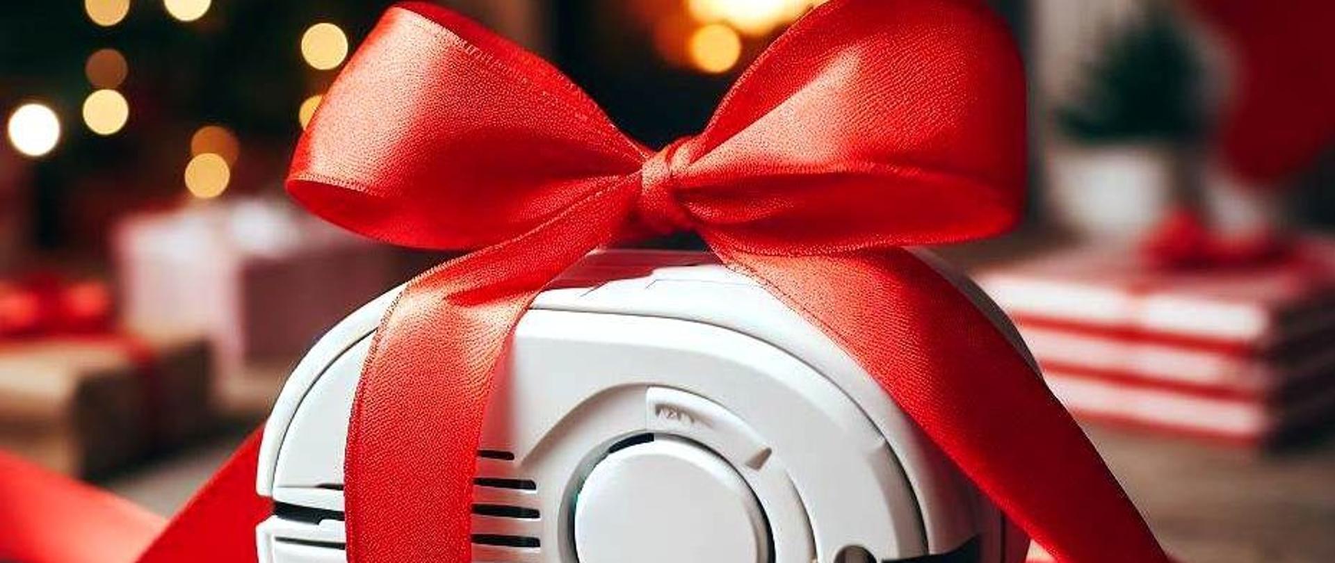 Na zdjęciu, na pierwszym planie pokazana jest czujka czadu lub dymu przewiązana szeroką czerwoną wstęgą. W tle światełka choinkowe oraz zapakowane prezenty.