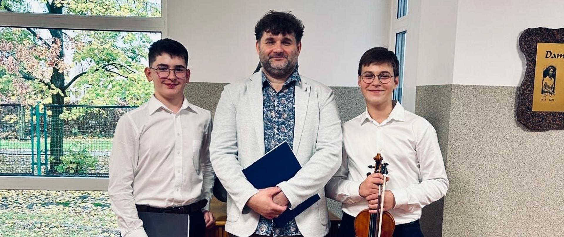 Na zdjęciu widać trzech mężczyzn stojących w pomieszczeniu o jasnych ścianach i dużym oknie, przez które widać jesienny krajobraz z drzewami. Wszyscy są elegancko ubrani — dwaj młodsi mężczyźni po bokach mają na sobie białe koszule i ciemne spodnie, a nauczyciel stojący w środku ma jasną marynarkę, wzorzystą koszulę i brązowe buty.
Osoba po prawej stronie trzyma skrzypce i smyczek, pozostałe dwie osoby trzymają w rękach granatowe teczki. W tle widać fragment tablicy pamiątkowej na ścianie oraz drewniany element wystroju wnętrza. 