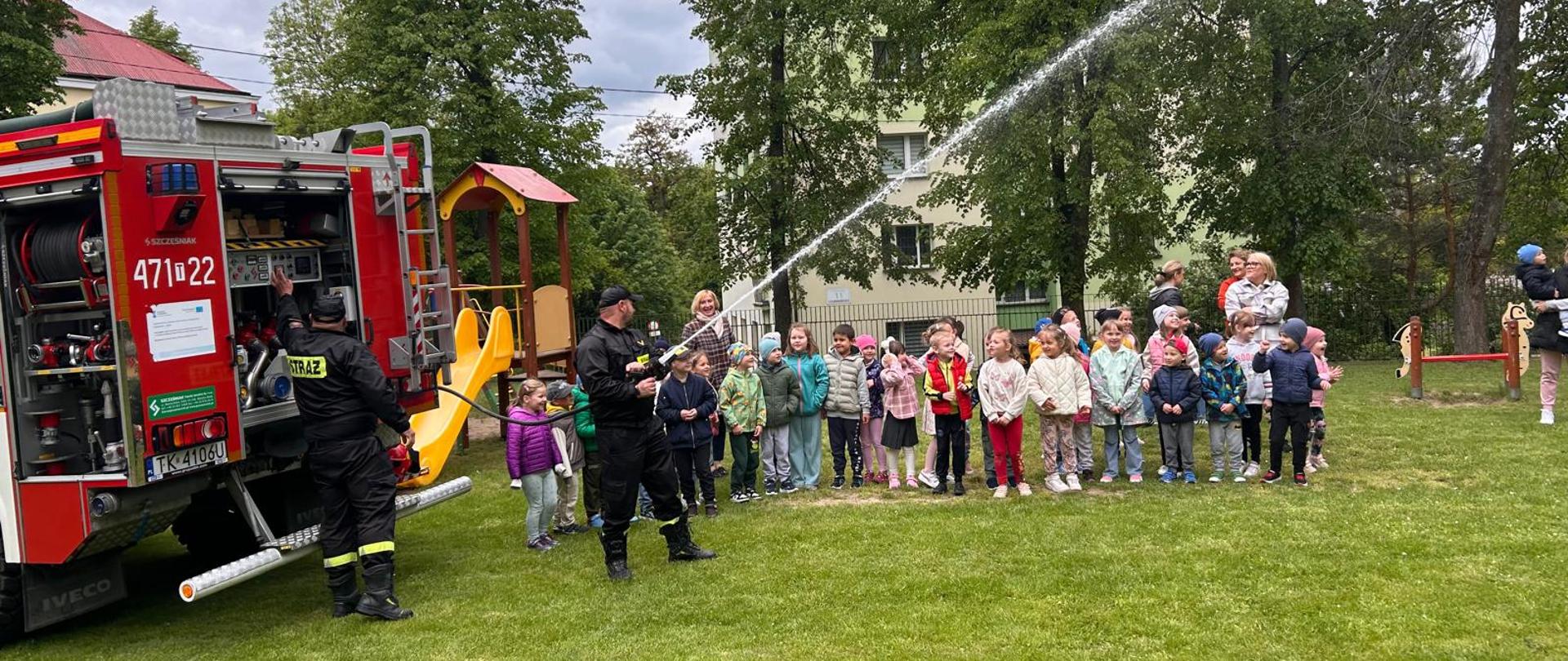 Strażacy wraz z przedszkolakami na tle wozu strażackiego.
