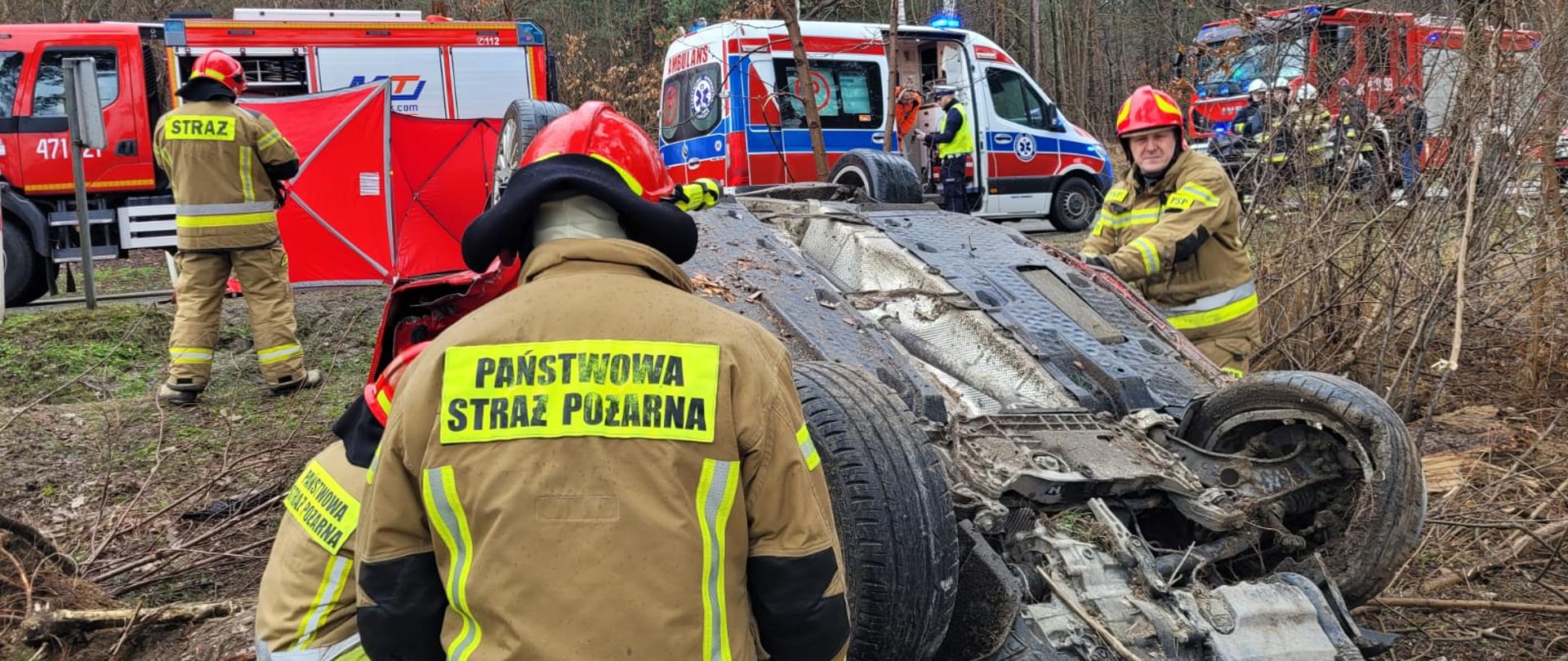 Samochód Pożarniczy, w tle dwa ambulans, samochód biorący udział w zdarzeniu a także strażaków pracujących na miejscu zdarzenia.