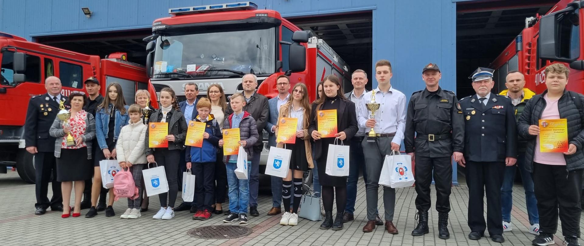 Zdjęcie przedstawia wszystkich laureatów Ogólnopolskiego Turnieju Wiedzy Pożarniczej „Młodzież zapobiega pożarom” wraz z opiekunami. Wszyscy znajdują się przed bramami garażowymi na tle samochodów ratowniczo-gaśniczych. Na zdjęciu widnieje także starosta jędrzejowski Paweł Faryna, komendant powiatowy Państwowej Straży Pożarnej w Jędrzejowie mł. bryg. Mariusz Stolarczyk oraz prezes Zarządu Oddziału Powiatowego Związku OSP RP w Jędrzejowie dh Tadeusz Soboń.
