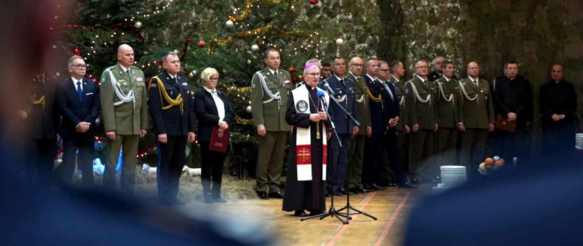 Zdjęcie zrobione w pomieszczeniu. Na zdjęciu uczestnicy Wigilii służb mundurowych. Na pierwszym planie przed mikrofonem stoi biskup rzeszowski.