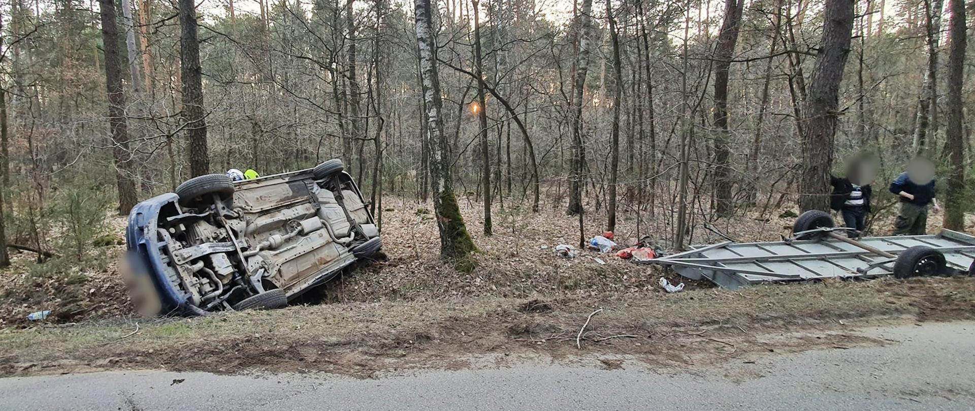 Na zdjęciu wrak samochodu osobowego i przyczepki po dachowaniu oraz strażacy udzielający pomocy.