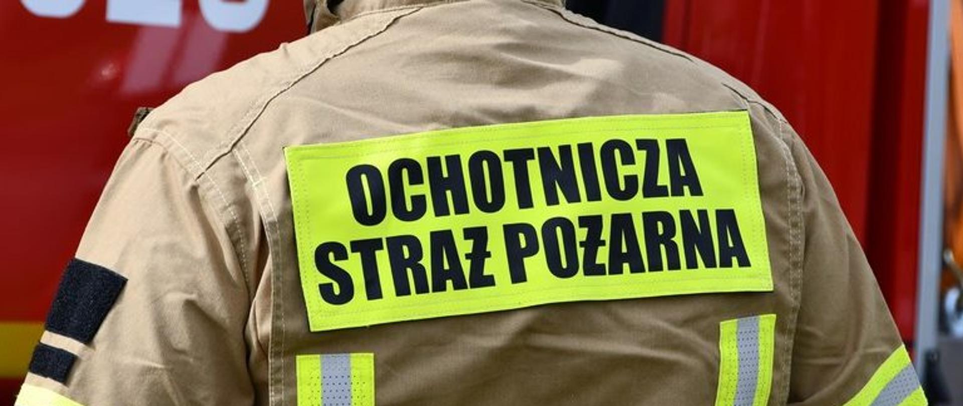 Plecy strażaka w mundurze bojowym z żółtą, fluorescencyjną naszywką z czarnym napisem: OCHOTNICZA STRAŻ POŻARNA