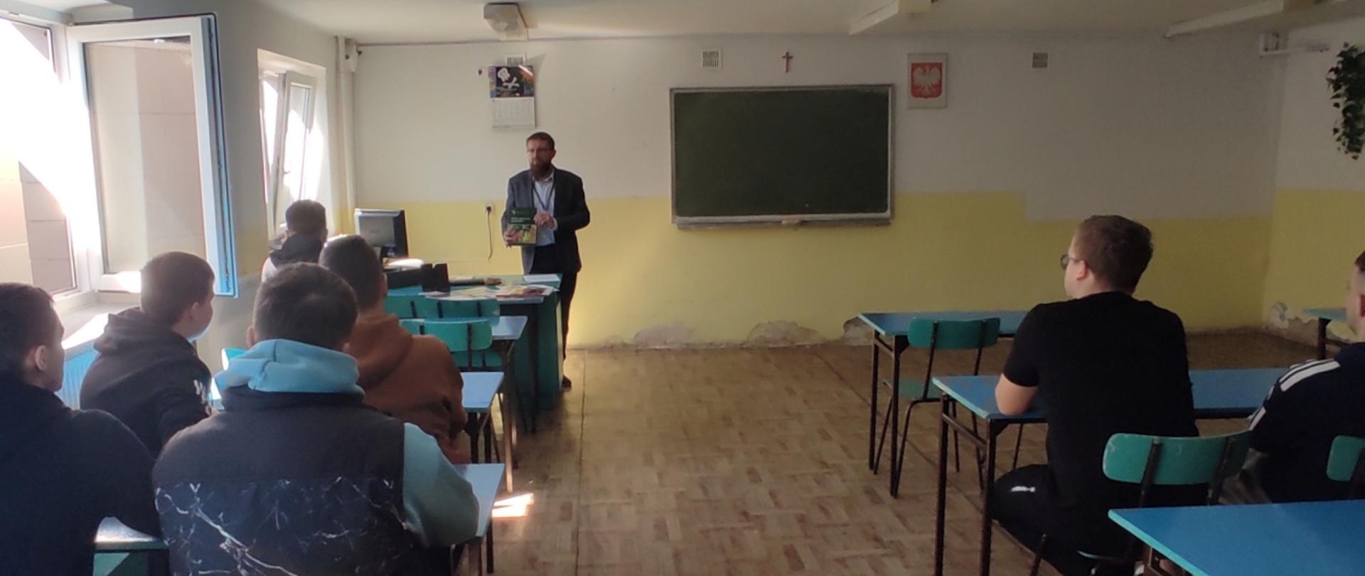 Zdjęcie przedstawia salę lekcyjną, w której prowadzone jest spotkanie. Kierownik Wojewódzkiego Inspektoratu ochrony Roślin i Nasiennictwa w Warszawie, Oddziału w Łosicach stoi przy tablicy i trzyma w rękach broszurę. Uczniowie lub uczestnicy siedzą w ławkach skierowanych w jego stronę.