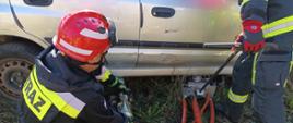 Strażak w ubraniu specjalnym kuca trzymając specjalistyczny sprzęt drugi za pomocą sprzętu podnosi samochód.