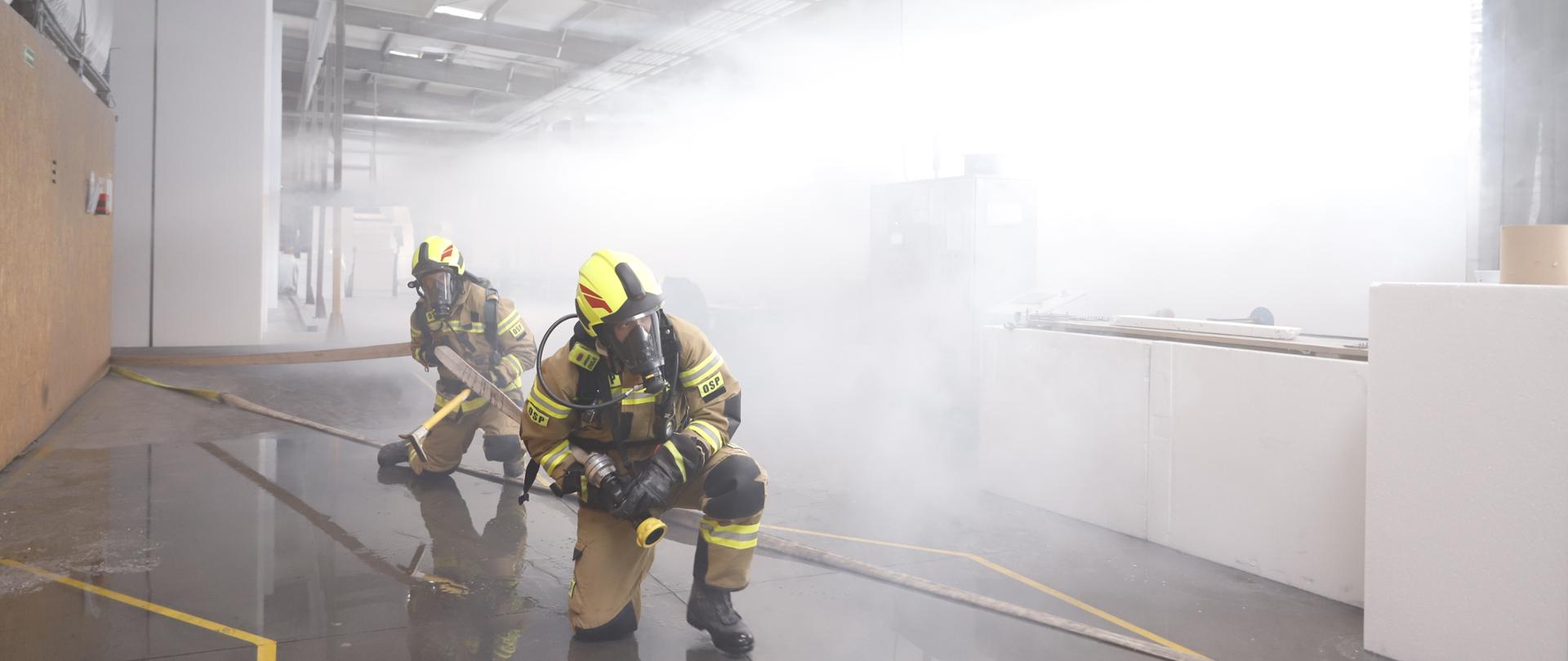 Strażacy poruszają się na terenie hali produkcyjnej, w tle zadymienie