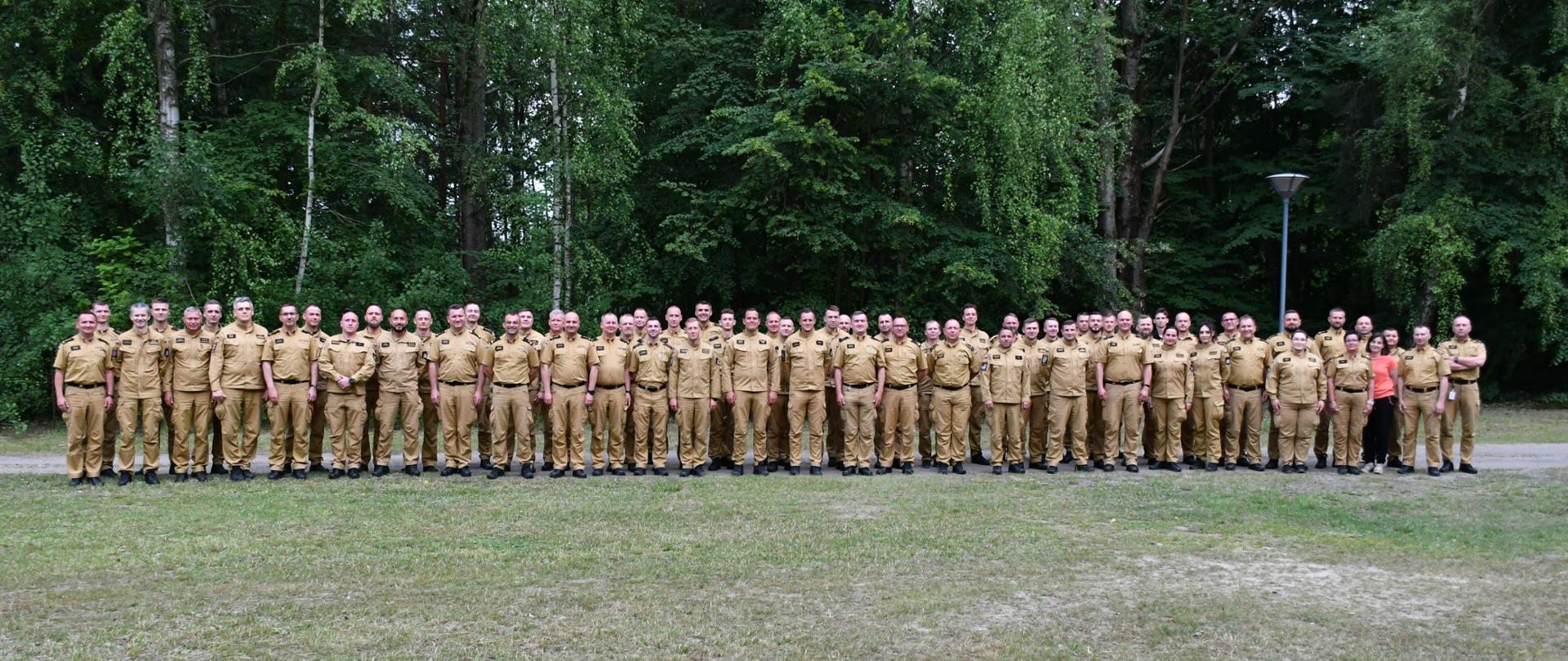 Zdjęcie grupowe kilkudziesięciu uczestników szkolenia stających na trawniku. W tle wysokie zielone drzewa. 