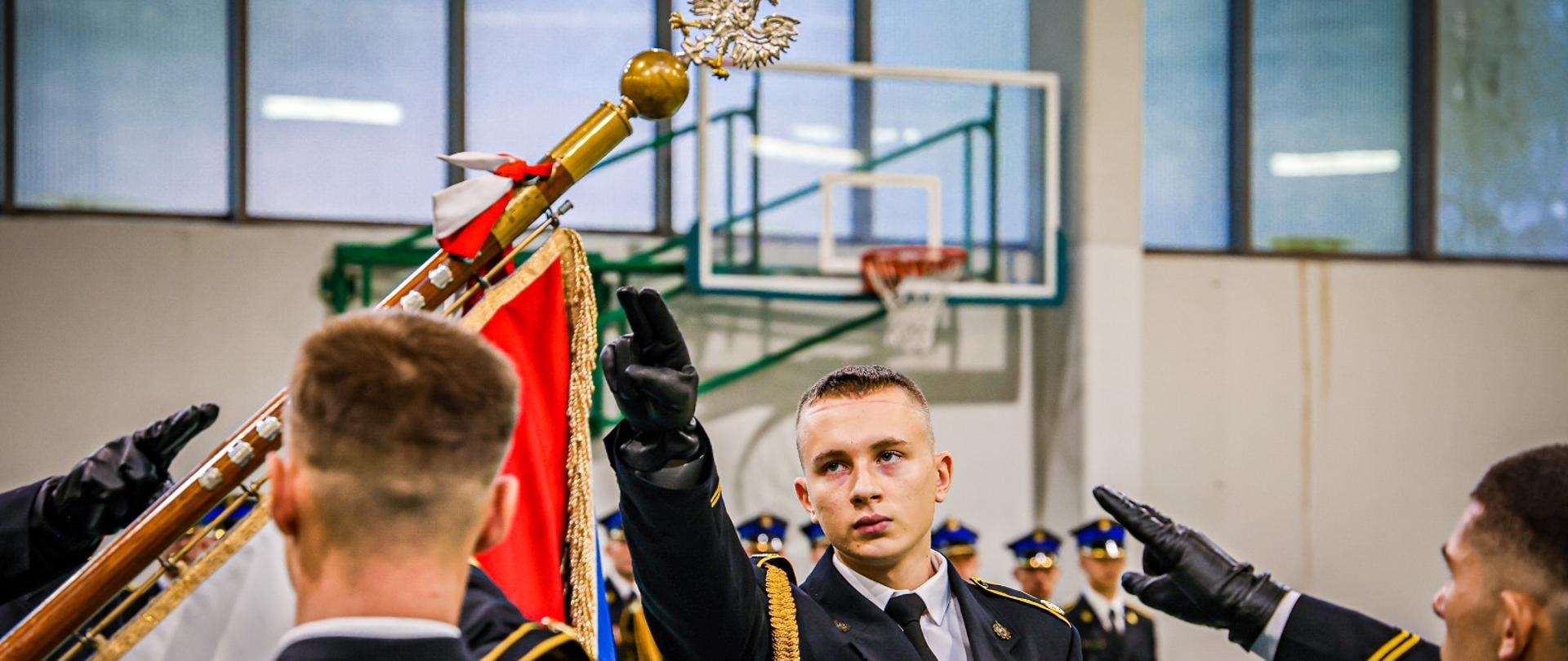Zdjęcie przedstawia uroczystość ślubowania w Szkole Aspirantów Państwowej Straży Pożarnej. Młody mężczyzna w czarnym, galowym mundurze strażackim PSP, z żółtymi naramiennikami i złotym sznurem, stoi na pierwszym planie i wykonuje gest unasząc rękę w stronę sztandaru. Obok niego, częściowo widoczny jest sztandar SAPSP . W tle widać innych słuchaczy w podobnych mundurach oraz elementy sali gimnastycznej.