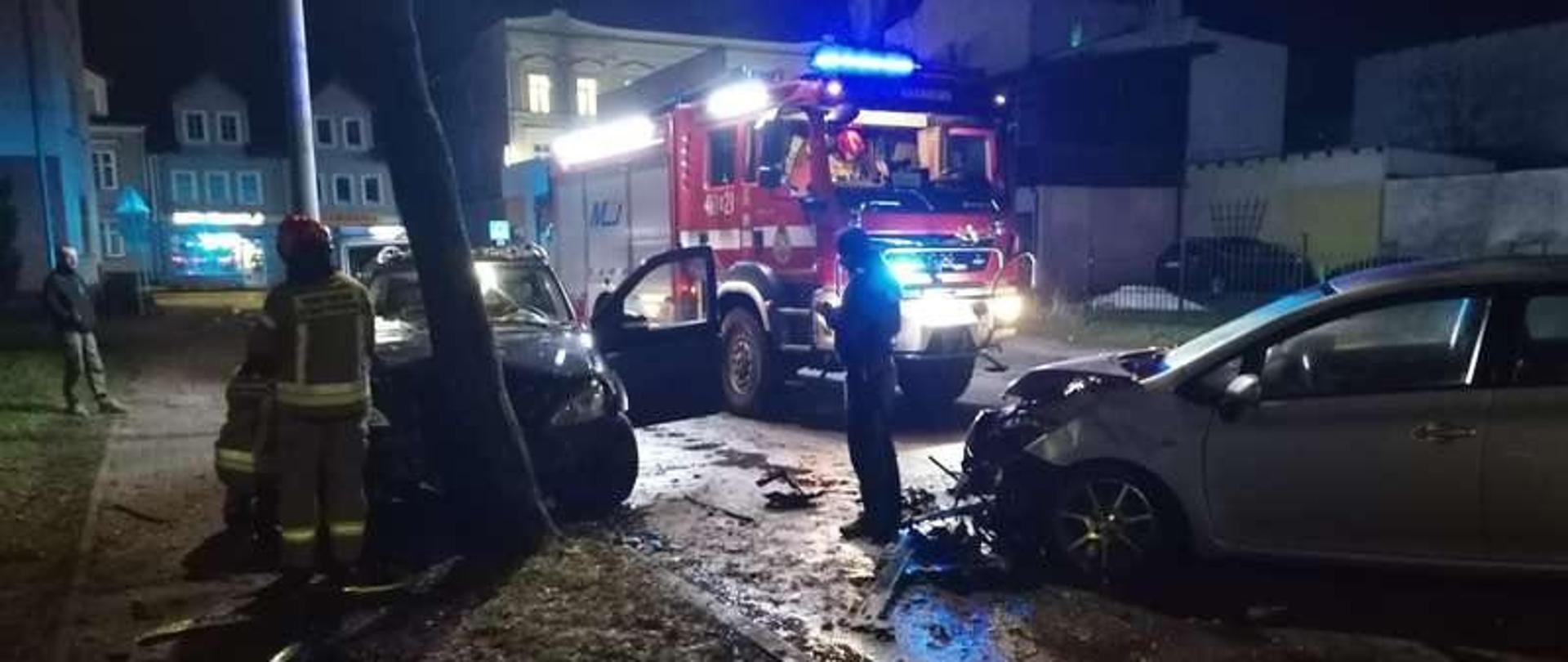 Zdjęcie zrobione nocą. Na drodze z prawej strony rozbite szare auto. Z lewej strony uszkodzone ciemne auto, rozbite o drzewo. Za nimi duży czerwony samochód strażacki na światłach ostrzegawczych, oświetla teren.